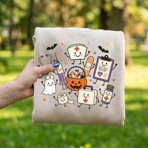 May include: A beige sweatshirt featuring a colorful graphic print of various medical-themed characters including a ghost, a syringe, a jack-o'-lantern, a bandage, a clipboard labeled BOO-BOO REPORT, a first aid kit, and a pill bottle with bats.