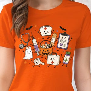 May include: An orange t-shirt featuring a graphic print of various medical-themed characters including a stethoscope, syringe, ghost, bandage, clipboard labeled BOO-BOO REPORT, pumpkin bucket, roll of tape, first aid kit, and medicine bottle.