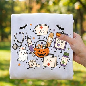 May include: A white sweatshirt featuring a colorful Halloween-themed medical graphic. The design includes a smiling ghost, a stethoscope, a syringe, a jack-o'-lantern candy bucket, a bandage, a first aid kit, and a clipboard labeled BOO-BOO REPORT.