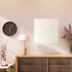 May include: A white textured canvas artwork hangs on a white wall above a wooden cabinet with a black metal mesh front. The cabinet is in a living room with a wooden floor, a clock, a lamp, and a vase of flowers.