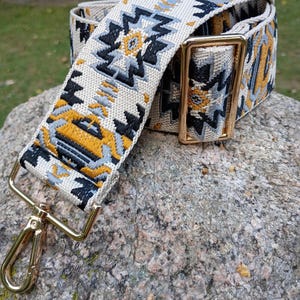 May include: A woven bag strap with a geometric pattern in cream, black, yellow, and gray. The strap has gold-colored metal hardware, including a clasp and a rectangular buckle. The strap is resting on a gray rock.