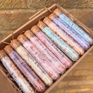 May include: A selection of scented bath salts in glass tubes with cork stoppers, presented in a brown cardboard box. Each tube is labelled with a different scent, such as Jasmine & Ylang Ylang, and Rosemary & Peppermint. The salts are various colours.