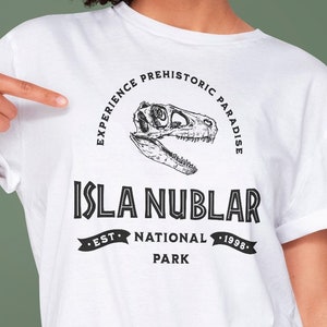 以下が含まれることがあります： 恐竜の頭蓋骨と「Experience Prehistoric Paradise Isla Nublar - Est. National Park - 1998.」というテキストが描かれた黒いグラフィックデザインが施された白いTシャツです。