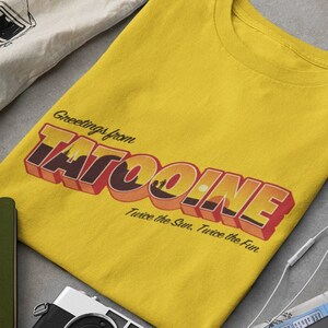 Op de afbeelding: Een geel T-shirt met een retro-grafische afbeelding met de tekst "Greetings from Tatooine. Twice the Sun. Twice the Fun."