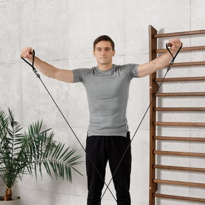 May include: A man wearing a grey t-shirt and black pants is using resistance bands to exercise. He is standing in front of a wooden wall with a ladder.