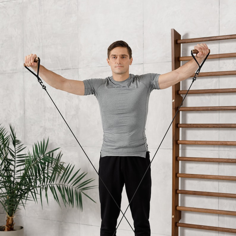 May include: A man wearing a grey t-shirt and black pants is using resistance bands to exercise. He is standing in front of a wooden wall with a ladder.