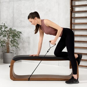 May include: A woman is using a resistance band while sitting on a wooden bench with a black leather seat. The bench has a curved design and is made of dark wood. The woman is wearing black leggings and a pink tank top.