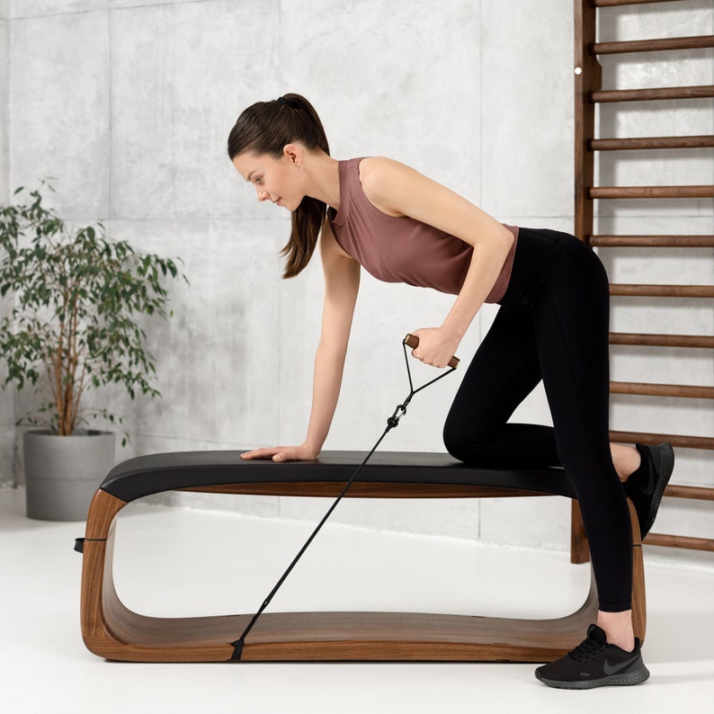 May include: A woman is using a resistance band while sitting on a wooden bench with a black leather seat. The bench has a curved design and is made of dark wood. The woman is wearing black leggings and a pink tank top.