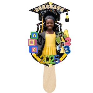 May include: Graduation photo prop featuring a young person in a yellow dress and graduation cap. The prop includes the word "CONGRATS", the year "2025", and colorful numbers and blocks. The prop is held by a wooden stick.