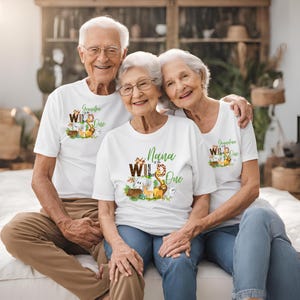 May include: Three people wearing white t-shirts with a jungle animal theme. The shirts say "Grandpa of the Wild One", "Nana of the Wild One", and "Grandmother of the Wild One".