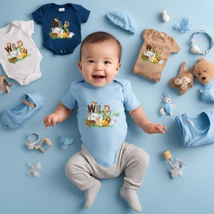 May include: A baby boy wearing a light blue bodysuit with the text 'Wild One' and a graphic of jungle animals. The baby is surrounded by various toys, including stuffed animals, a blue hat, and a white onesie with the text 'Wild' and a jungle animal graphic.