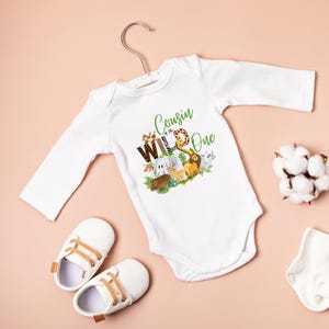 May include: White long-sleeved onesie with a green and brown graphic of animals and the text "Cousin of the Wild One".