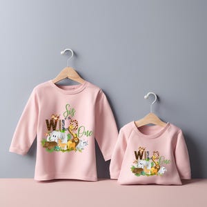 May include: Two pink long-sleeved shirts with a jungle animal graphic and the text "Sis of the Wild One" and "Wild One".