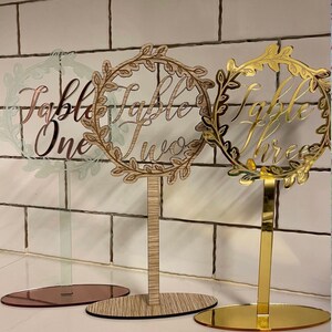 May include: Three table number signs with a wreath design. The signs are made of clear acrylic with gold, wood, and rose gold bases. The signs read "Table One", "Table Two", and "Table Three".