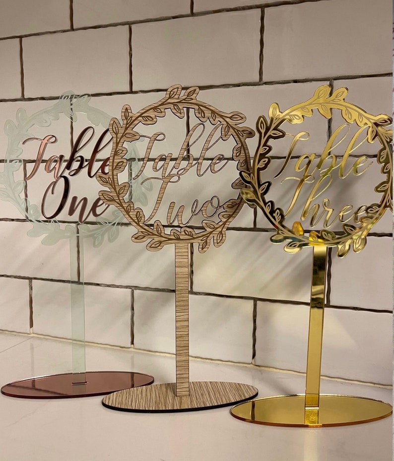 May include: Three table number signs with a wreath design. The signs are made of clear acrylic with gold, wood, and rose gold bases. The signs read "Table One", "Table Two", and "Table Three".