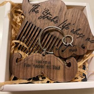 May include: A wooden box with a mustache-shaped comb and keychain. The comb has the text "The Stache Life" engraved on it. The keychain has a mustache shape and the text "You're a Fan 'tache' tic Dad" engraved on it. The keychain also has a smaller mustache with the name "Paul" engraved on it.