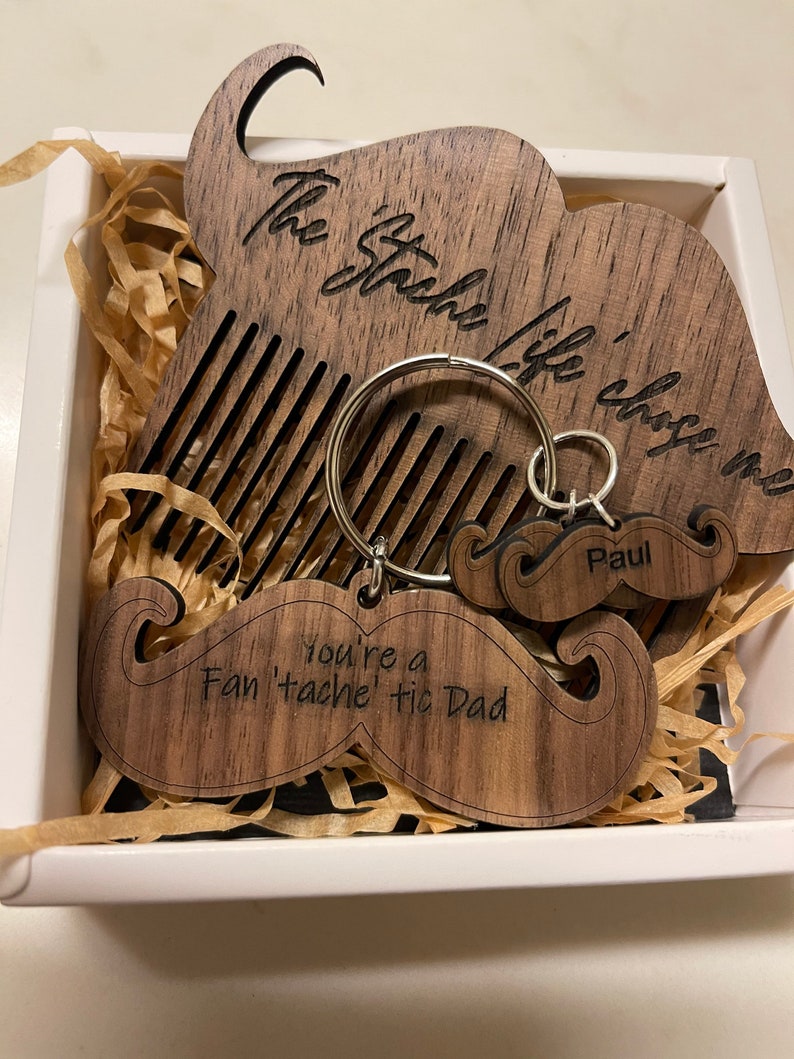 May include: A wooden box with a mustache-shaped comb and keychain. The comb has the text "The Stache Life" engraved on it. The keychain has a mustache shape and the text "You're a Fan 'tache' tic Dad" engraved on it. The keychain also has a smaller mustache with the name "Paul" engraved on it.