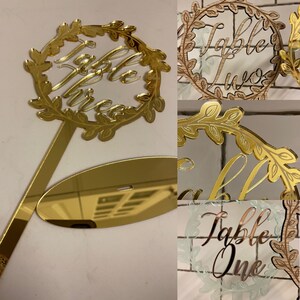 May include: Gold mirror acrylic table number with a leafy wreath design. The number three is written in cursive inside the wreath. The table number comes with a separate stand.