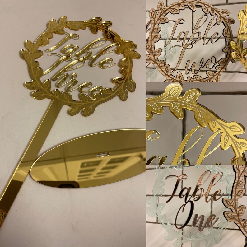 May include: Gold mirror acrylic table number with a leafy wreath design. The number three is written in cursive inside the wreath. The table number comes with a separate stand.