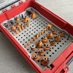 May include: A red toolbox with a gray interior, filled with a variety of orange and silver router bits. The bits are arranged in a grid pattern, showcasing different shapes and sizes for woodworking projects.
