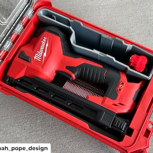 May include: A red and black Milwaukee power tool is stored in a red hard case with a clear lid. The tool is a cordless nail gun with a black handle and a red body. The case has a foam insert to protect the tool.
