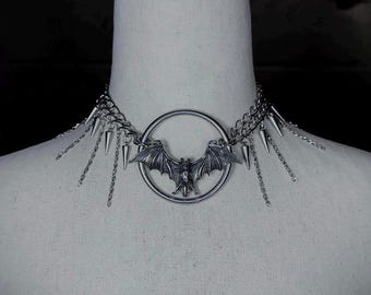 May include: A silver choker necklace with a bat pendant and spikes. The necklace is on a white mannequin.