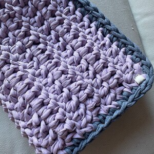 Puede incluir: Primer plano de una manta de crochet de color morado y gris. La manta est&aacute; hecha con un hilo grueso y tiene una superficie texturizada.