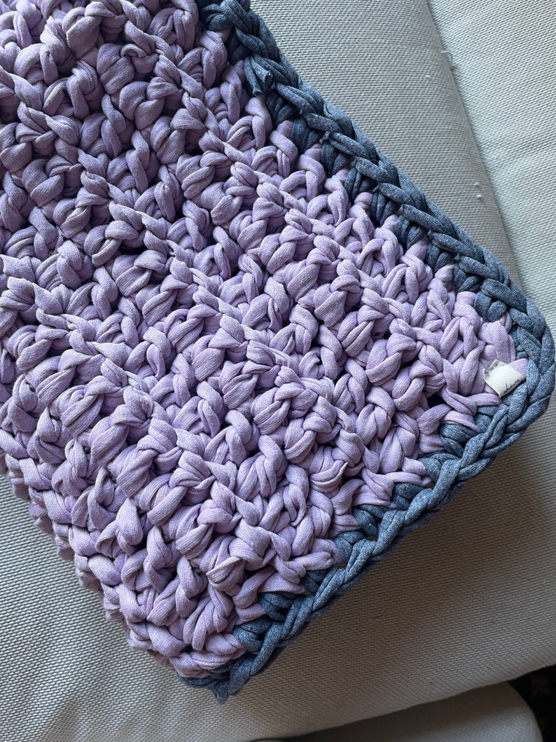 Puede incluir: Primer plano de una manta de crochet de color morado y gris. La manta est&aacute; hecha con un hilo grueso y tiene una superficie texturizada.