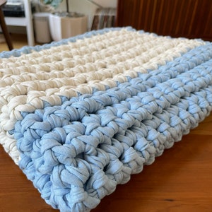 Puede incluir: Una almohada de punto de crochet con un patr&oacute;n de rayas azul claro y blanco. La almohada est&aacute; hecha de un hilo suave y texturizado y tiene un dise&ntilde;o simple y moderno.