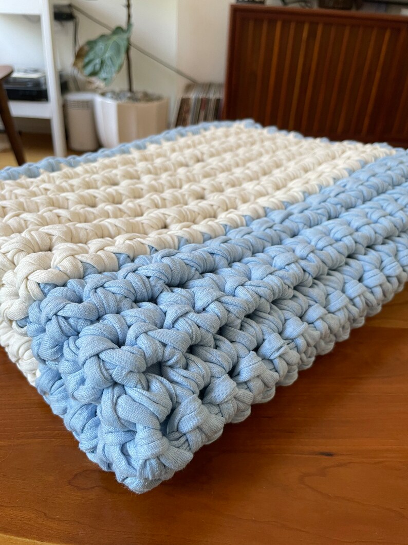 Puede incluir: Una almohada de punto de crochet con un patr&oacute;n de rayas azul claro y blanco. La almohada est&aacute; hecha de un hilo suave y texturizado y tiene un dise&ntilde;o simple y moderno.