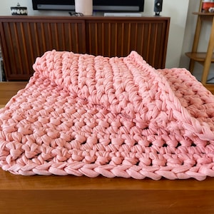 Puede incluir: Una manta de crochet rosa hecha de tiras de tela. La manta est&aacute; doblada por la mitad y est&aacute; sobre una superficie de madera.