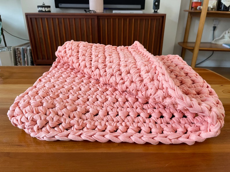 Puede incluir: Una manta de crochet rosa hecha de tiras de tela. La manta est&aacute; doblada por la mitad y est&aacute; sobre una superficie de madera.