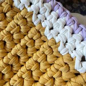 Puede incluir: Primer plano de un coj&iacute;n tejido a crochet con un esquema de colores amarillo, blanco y morado. El coj&iacute;n est&aacute; hecho de hilo grueso y tiene una superficie texturizada.