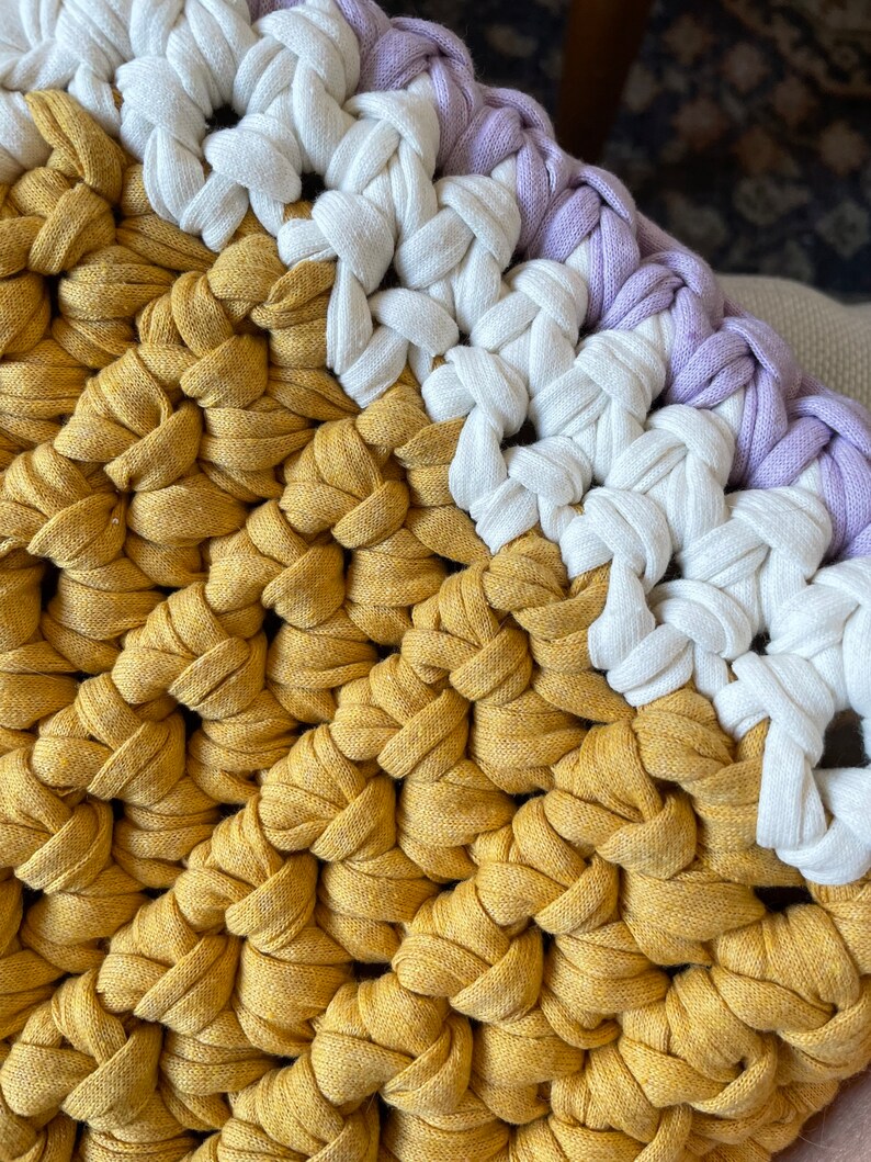 Puede incluir: Primer plano de un coj&iacute;n tejido a crochet con un esquema de colores amarillo, blanco y morado. El coj&iacute;n est&aacute; hecho de hilo grueso y tiene una superficie texturizada.