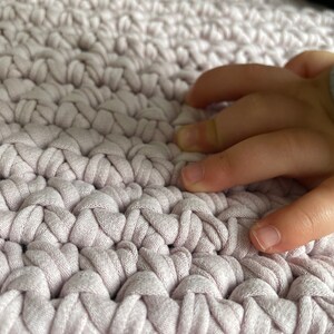 Puede incluir: Primer plano de una alfombra de crochet rosa claro con una mano descansando sobre la superficie.
