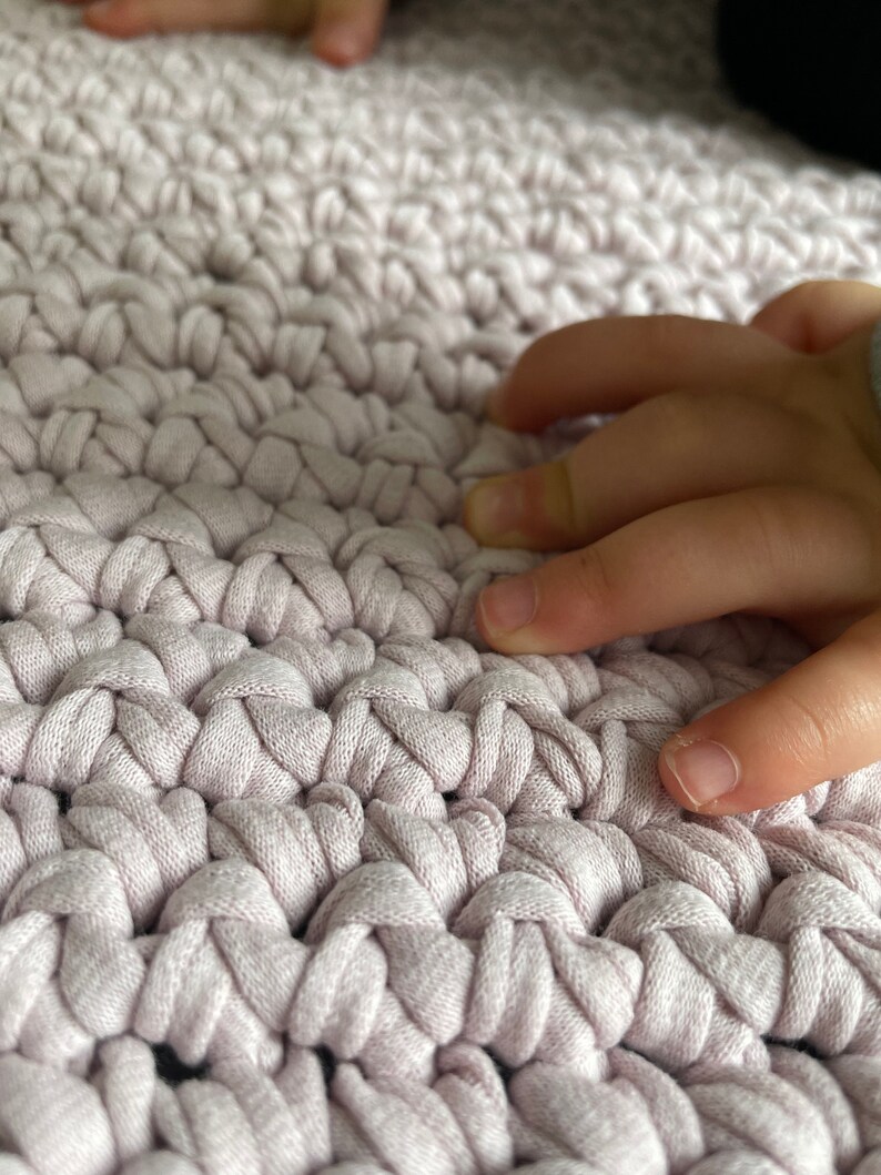 Puede incluir: Primer plano de una alfombra de crochet rosa claro con una mano descansando sobre la superficie.