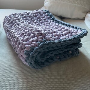Puede incluir: Una manta de ganchillo doblada con un esquema de color morado y gris. La manta est&aacute; hecha con un hilo grueso y tiene un patr&oacute;n de punto simple.