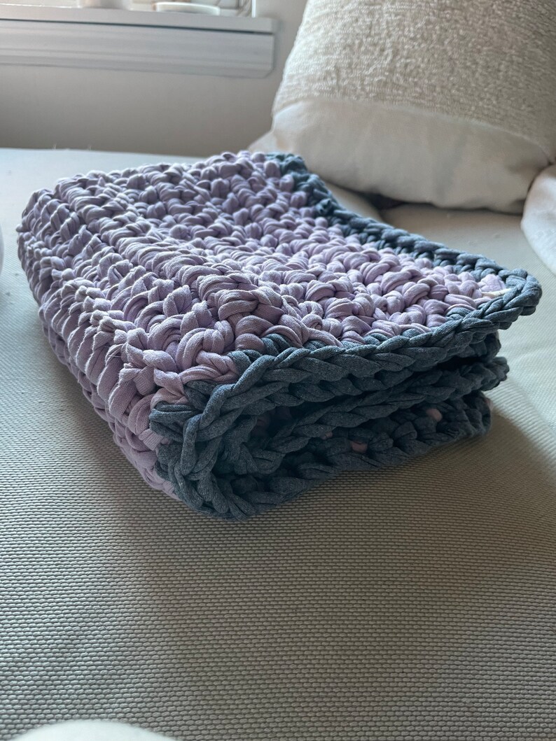 Puede incluir: Una manta de ganchillo doblada con un esquema de color morado y gris. La manta est&aacute; hecha con un hilo grueso y tiene un patr&oacute;n de punto simple.