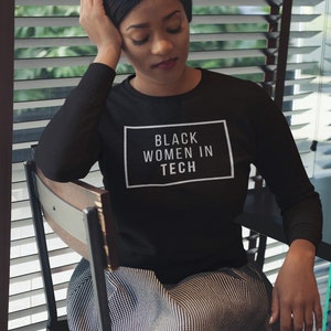 May include: A black long-sleeved sweatshirt with the text "BLACK WOMEN IN TECH" in a white rectangular box. The person is wearing black and white checkered pants and is sitting in a chair.