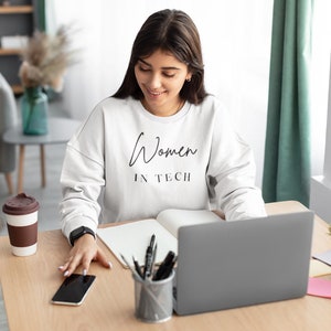 May include: A woman wearing a white sweatshirt with the text "Women in Tech" printed on it. She is sitting at a desk with a laptop, a notebook, a cup of coffee, and a smartphone.