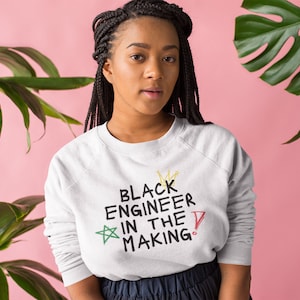 Könnte beinhalten: Ein weißer Pullover mit dem Text "Black Engineer in the Making" in schwarzer Schrift. Der Text ist von einem Stern, einer Krone und einem Ausrufezeichen umgeben.