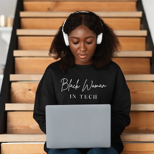 Puede incluir: Una mujer negra con auriculares está sentada en una escalera de madera y usa una computadora portátil. La computadora portátil está abierta y la pantalla es visible. La mujer lleva una sudadera negra con el texto "Black Woman in Tech" impreso en la parte delantera.