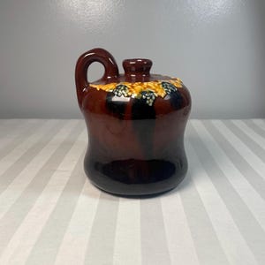 May include: A brown ceramic jug with a handle and a lid. The jug has a dark brown base that fades to a lighter brown with a decorative band of yellow and blue grapes around the top. The jug is on a striped surface.