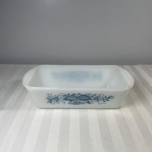 May include: A rectangular, white glass baking dish with rounded corners. It has a teal floral pattern around the sides. The interior is empty, and the dish rests on a striped surface.