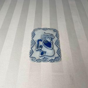 May include: A rectangular, white ceramic wall hanging with a blue angel design. The angel is depicted with wings and a draped garment. The border features a detailed, blue, scalloped pattern. A small hole is at the top for hanging.