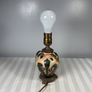 May include: A small, antique table lamp with a cream-colored ceramic base featuring green and red floral designs. The lamp has a brass fixture and a white bulb. The base sits on a decorative, bronze-colored metal stand. A brown electrical cord extends from the base.