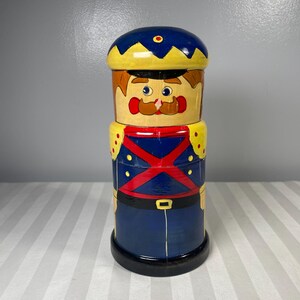 May include: A hand-painted wooden stacking doll in the shape of a soldier. The doll features a blue hat, a yellow and red uniform, and a black base. The doll has a painted face with a mustache and a stern expression.