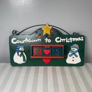 May include: A green wooden Christmas countdown sign with the words "Countdown to Christmas" in white. The sign features two snowmen, a star, and a heart. The countdown shows "2" and "5".