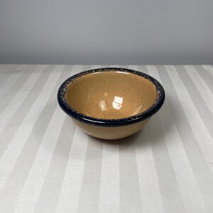 Może przedstawiać: Mała, okrągła ceramiczna miska z beżowym wnętrzem i ciemnoniebieskim obramowaniem. Misa ma nakrapiany wygląd i prawdopodobnie służy do serwowania jedzenia lub jako przedmiot dekoracyjny. Misa stoi na biało-szarej powierzchni w paski.