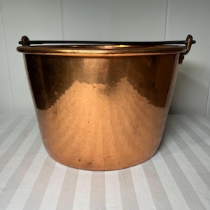 Puede incluir: Un cubo de cobre con textura martillada y un asa de metal negro. El cubo tiene una abertura ancha y una base redondeada. El cobre tiene un color marrón rojizo cálido.